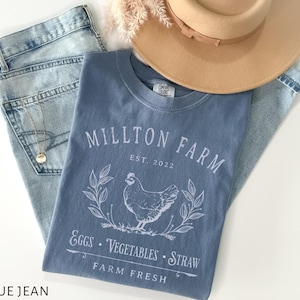 Puede incluir: Una camiseta de mezclilla azul con un dise&ntilde;o gr&aacute;fico blanco que dice "Millton Farm Est. 2022" con una ilustraci&oacute;n blanca de un pollo y las palabras "Eggs, Vegetables, Straw Farm Fresh".