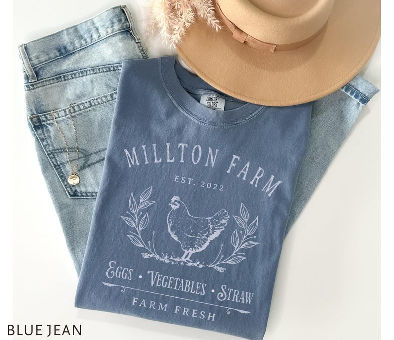 Puede incluir: Una camiseta de mezclilla azul con un dise&ntilde;o gr&aacute;fico blanco que dice "Millton Farm Est. 2022" con una ilustraci&oacute;n blanca de un pollo y las palabras "Eggs, Vegetables, Straw Farm Fresh".
