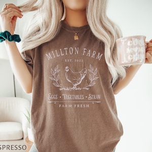Puede incluir: Una camiseta marr&oacute;n con el texto "Millton Farm Est. 2022" y una ilustraci&oacute;n blanca de una gallina, huevos, verduras y paja. El texto "Farm Fresh" est&aacute; debajo de la ilustraci&oacute;n.