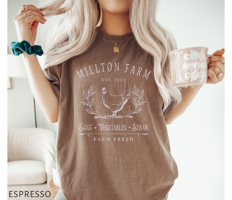 Puede incluir: Una camiseta marr&oacute;n con el texto "Millton Farm Est. 2022" y una ilustraci&oacute;n blanca de una gallina, huevos, verduras y paja. El texto "Farm Fresh" est&aacute; debajo de la ilustraci&oacute;n.