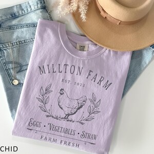 Puede incluir: Una camiseta lavanda con el texto "Millton Farm Est. 2022" y una ilustraci&oacute;n en blanco y negro de una gallina, hojas y el texto "Eggs - Vegetables - Straw Farm Fresh".