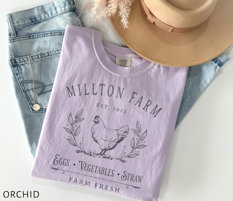 Puede incluir: Una camiseta lavanda con el texto "Millton Farm Est. 2022" y una ilustraci&oacute;n en blanco y negro de una gallina, hojas y el texto "Eggs - Vegetables - Straw Farm Fresh".