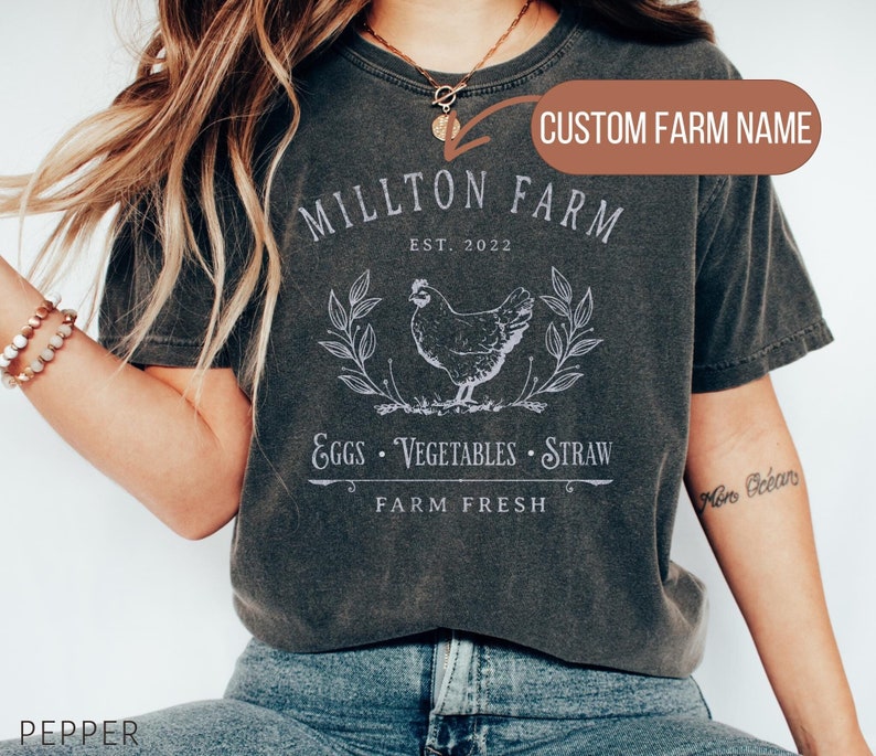 Puede incluir: Una camiseta gris oscuro con un dise&ntilde;o gr&aacute;fico blanco que presenta una gallina, hojas y el texto "MILLTON FARM EST. 2022 EGGS VEGETABLES STRAW FARM FRESH". El texto "CUSTOM FARM NAME" est&aacute; escrito en un cuadro marr&oacute;n sobre el gr&aacute;fico.