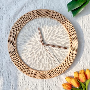 May include: A round, woven wicker wall clock with a white face and a gray floral pattern. The clock has brown hands.