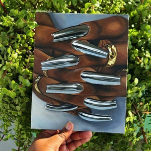 Op de afbeelding: Een schilderij van een hand met lange, zilveren chromen nagels en gouden ringen. Het kunstwerk wordt tegen een achtergrond van groen gebladerte gehouden. De Instagram-handle van de kunstenaar, @neonpalette.co, is linksboven zichtbaar.