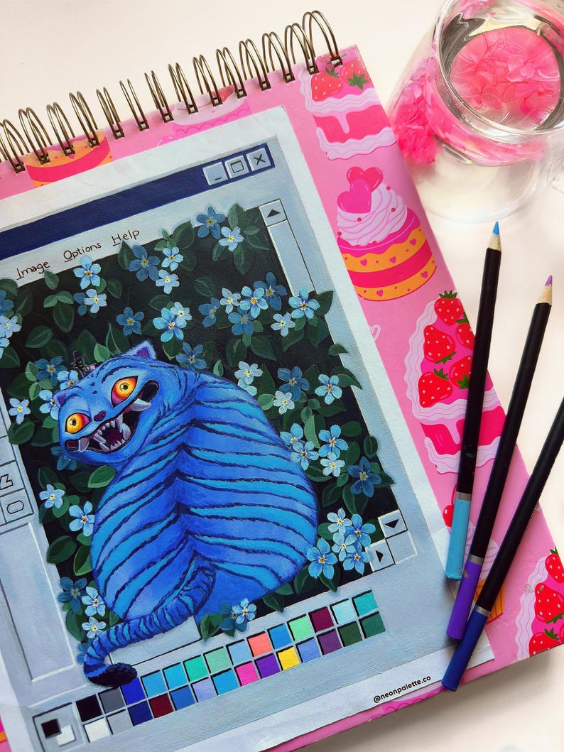 May include: A spiral-bound sketchbook with a pink cover featuring a whimsical cake design. The open page displays a vibrant illustration of a blue tiger within a computer window frame, surrounded by blue flowers. Colored pencils and a glass of water are also present.
