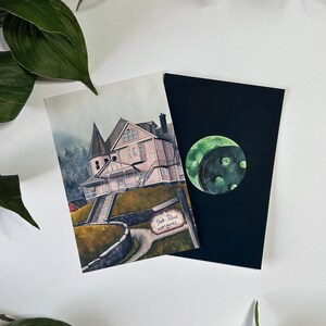 May include: A watercolor illustration of a pink house with a sign that reads "Pink Place Apartments". The house is on a hill with a stone wall and a path leading up to it. The illustration is on a white background. There is a separate black card with a green moon illustration.