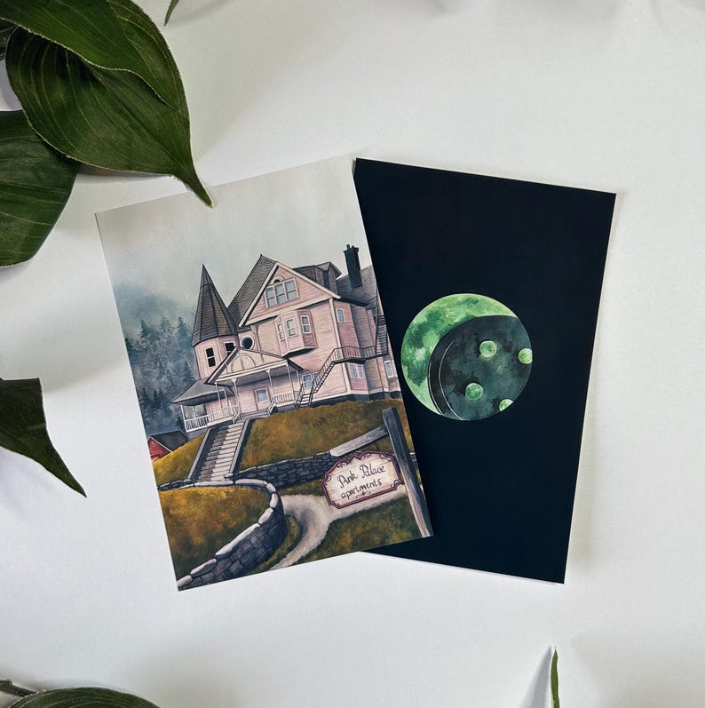 May include: A watercolor illustration of a pink house with a sign that reads "Pink Place Apartments". The house is on a hill with a stone wall and a path leading up to it. The illustration is on a white background. There is a separate black card with a green moon illustration.