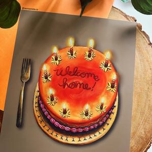 May include: A greeting card featuring a painted birthday cake with lit candles and the words "Welcome home!" in red script. A gold fork is placed to the left of the cake. The card is on a wooden surface.