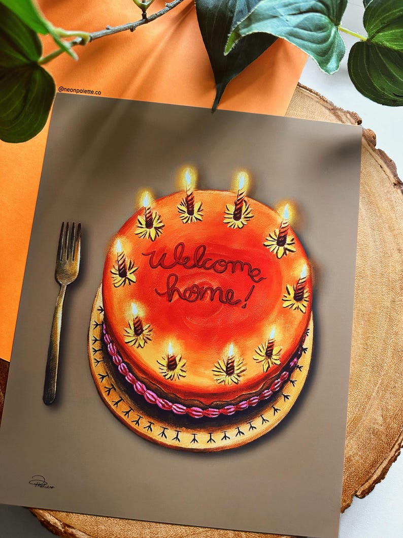 May include: A greeting card featuring a painted birthday cake with lit candles and the words "Welcome home!" in red script. A gold fork is placed to the left of the cake. The card is on a wooden surface.
