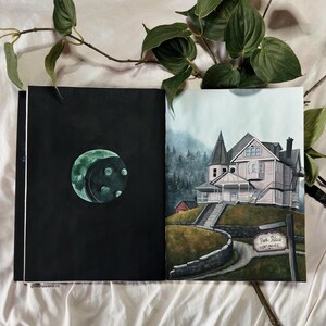 May include: A hand-painted illustration of a pink house with a sign that reads "Pink Palace Apartments". The house is surrounded by trees and a stone wall. The illustration is on the right page of an open sketchbook. The left page of the sketchbook is black with a green moon.