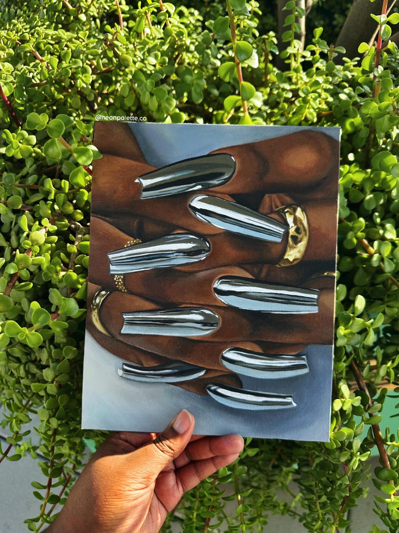 Op de afbeelding: Een schilderij van een hand met lange, zilveren chromen nagels en gouden ringen. Het kunstwerk wordt tegen een achtergrond van groen gebladerte gehouden. De Instagram-handle van de kunstenaar, @neonpalette.co, is linksboven zichtbaar.