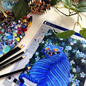 May include: An artistic still life featuring colored pencils, a paint palette, and a drawing of a blue cat with a menacing expression. The drawing is surrounded by blue flowers. The text "SAJA - Paint" is visible.