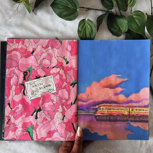 May include: An open sketchbook with two pages showing hand-painted illustrations. The left page features a pink floral pattern with a small white label that says "そうじゃないよ  君を好きだよ". The right page shows a blue sky with a pink cloud and a yellow train reflected in a body of water.