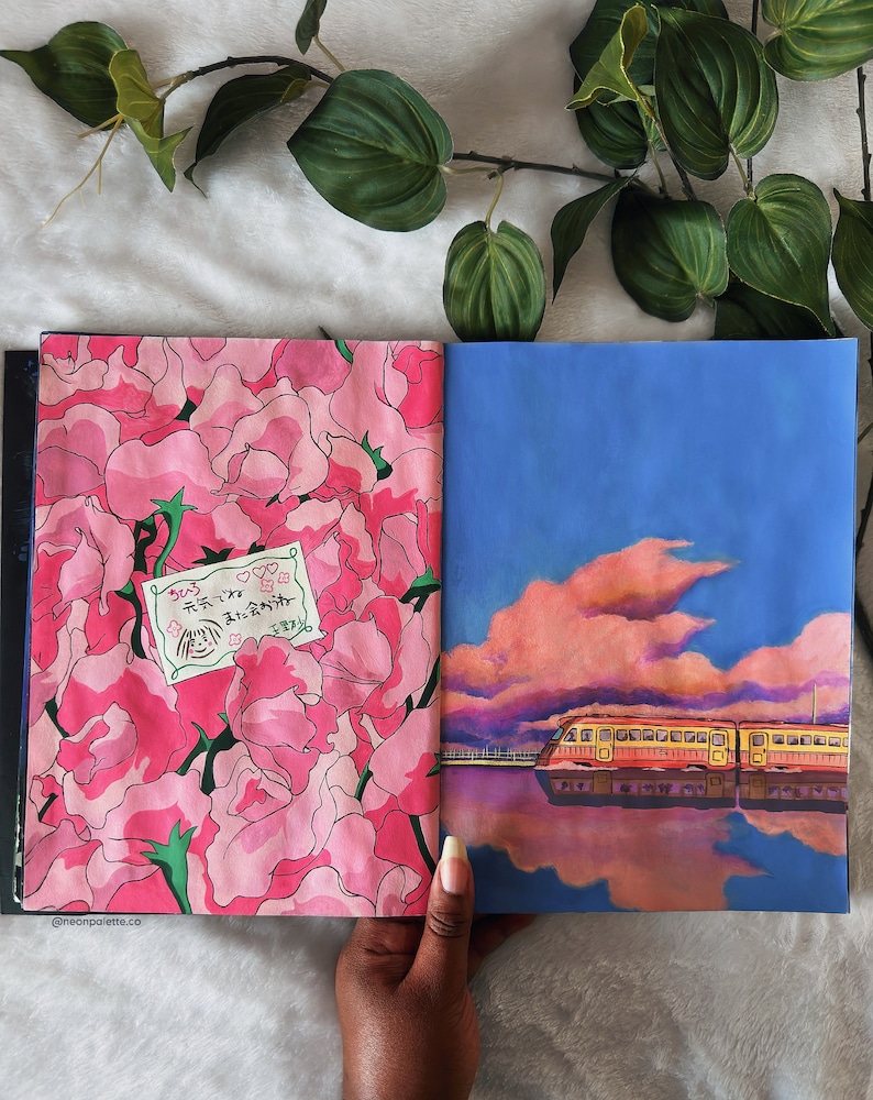 May include: An open sketchbook with two pages showing hand-painted illustrations. The left page features a pink floral pattern with a small white label that says "そうじゃないよ  君を好きだよ". The right page shows a blue sky with a pink cloud and a yellow train reflected in a body of water.