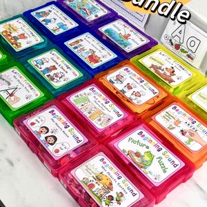 May include: A selection of brightly coloured task boxes labelled "Beginning Sound Task Box Bundle." The boxes are in various colours, including green, blue, pink, and orange. Each box contains educational materials for early learning.