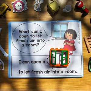 May include: A children's book page with the text "What can I open to let fresh air into a room?" and "I can open a to let fresh air into a room." A small girl in a red dress stands near a window illustration. Miniature toys surround the book.