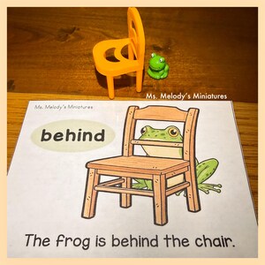 May include: An orange miniature chair and a small green frog figurine are positioned above a picture card. The card features a cartoon frog behind a wooden chair, with the word "behind" and the sentence "The frog is behind the chair."