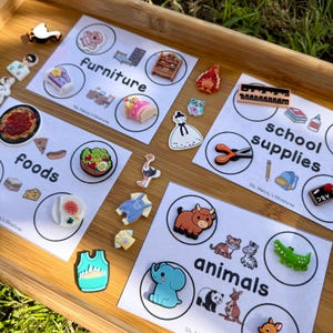 Puede incluir: Una bandeja de madera muestra tarjetas con objetos temáticos en miniatura. Las categorías incluyen "muebles", "alimentos", "útiles escolares" y "animales". Las miniaturas son coloridas y detalladas, incluyendo una cama, pizza, tijeras y un elefante.