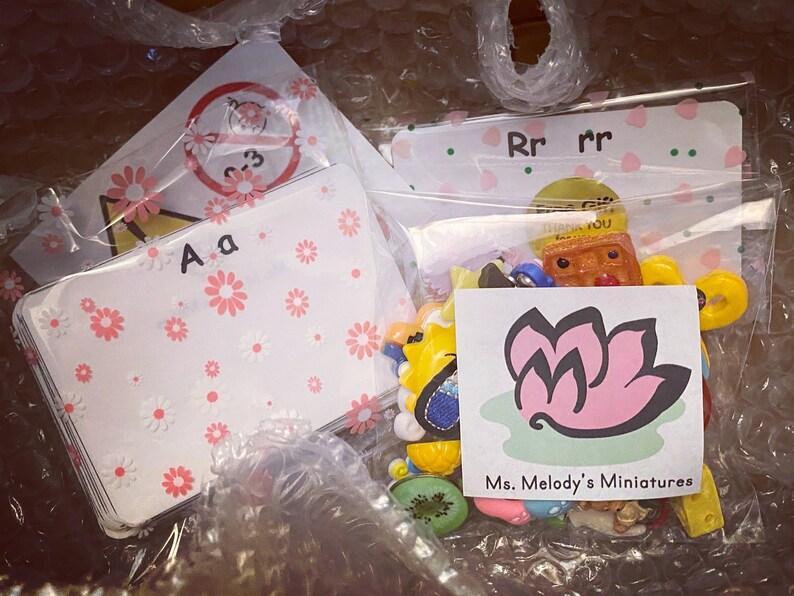 May include: A clear plastic bag filled with colorful miniature toys and trinkets. The bag also contains several white cards with pink flowers and letters printed on them. The cards say "Aa" and "Rr". A small white card with a pink flower logo and the text "Ms. Melody's Miniatures" is also visible.