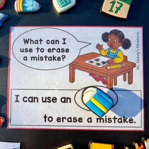 May include: A rectangular educational card featuring a cartoon of a child at a desk with a speech bubble asking, What can I use to erase a mistake? Below, the text reads, I can use an to erase a mistake, with a blue, yellow, and white eraser.