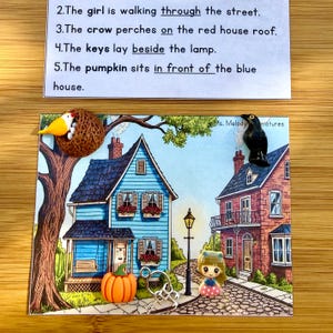 May include: A miniature scene with a blue house, a red house, a pumpkin, and a doll. A crow and a bird are perched on the houses. The text on the top says: "The girl is walking through the street."