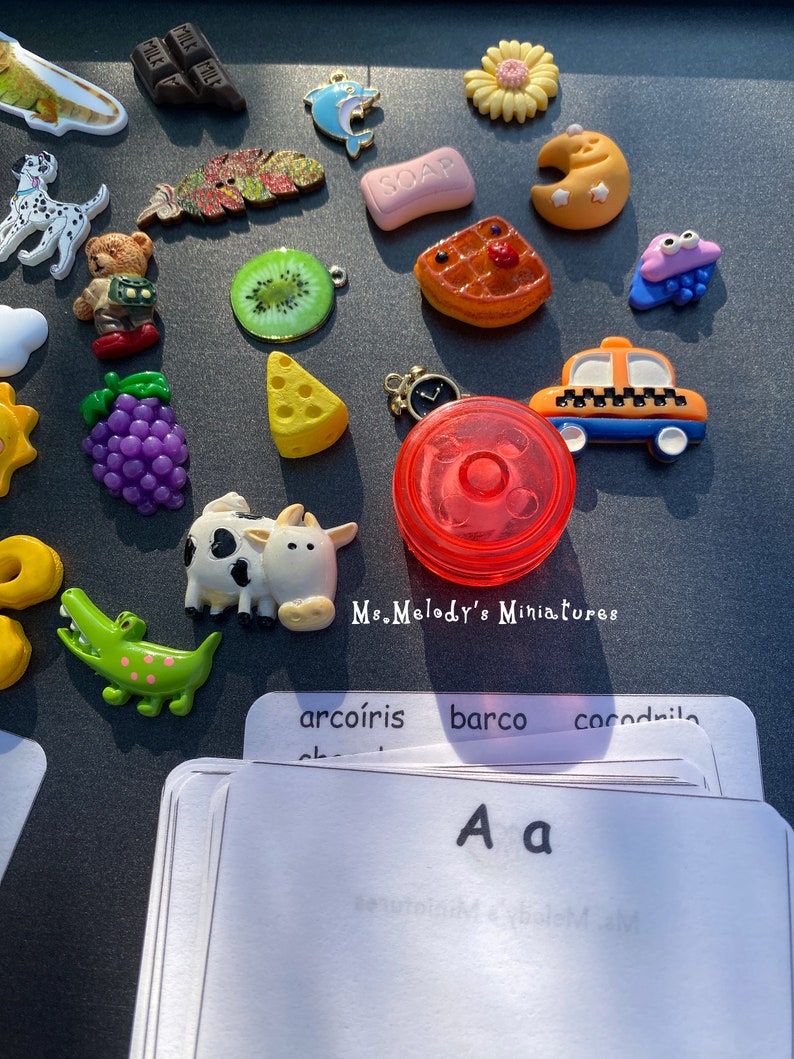 May include: A collection of colorful miniature toys, including a cow, a crocodile, a kiwi, a grape bunch, a yellow star, a red and white checkered pizza slice, a yellow taxi, a red spinning top, a blue dolphin, a green leaf, a bar of chocolate, a pink bar of soap, a yellow crescent moon, a purple monster with googly eyes, a white dog, a brown teddy bear, a white cloud, a yellow triangle, and a set of flashcards with the letter "A" written on them. The flashcards also have the words "arcoiris", "barco", and "cocodrilo" written on them.