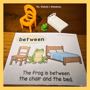 May include: An orange plastic chair, a green frog figurine, and a white miniature bed are displayed above a card. The card reads "between" and features a frog between a chair and a bed. The text on the card says, "The frog is between the chair and the bed."