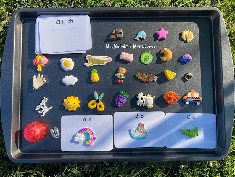 May include: A black baking sheet with white cards labeled 'CH ch', 'Aa', 'Bb', and 'Cc'. The cards have colorful miniature figurines on them, including a rainbow, a boat, a crocodile, a sun, a pineapple, a cloud, a hand, a dog, a bear, a cow, a kiwi, a crescent moon, a star, a flower, a dolphin, a piece of cheese, a feather, a pair of scissors, a bunch of grapes, a waffle, a taxi, an egg, a lizard, and a lollipop.