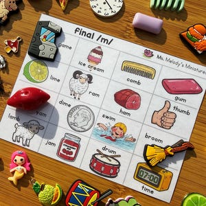 Puede incluir: Un colorido juego educativo con el texto "final /m/" y varios amuletos en miniatura. Los amuletos incluyen una consola de juegos, helado, un peine, un trozo de jamón, una moneda de diez centavos, un cordero y un tambor.
