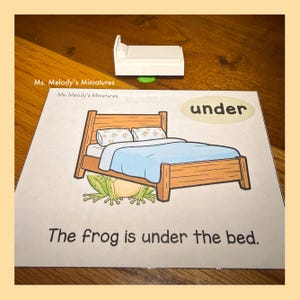 May include: A miniature white bed sits above a picture of a bed with a frog underneath. The picture has the words "under" and "The frog is under the bed." The bed in the picture is brown with a blue blanket and two pillows.