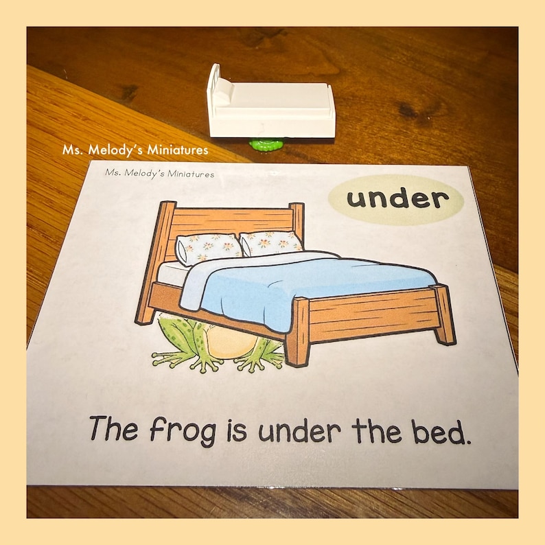 May include: A miniature white bed sits above a picture of a bed with a frog underneath. The picture has the words "under" and "The frog is under the bed." The bed in the picture is brown with a blue blanket and two pillows.