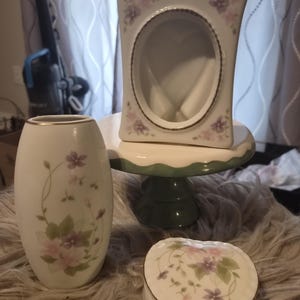 Puede incluir: Colección de objetos decorativos de porcelana. Incluye un jarrón blanco con detalles florales, una caja de baratijas en forma de corazón y un marco de fotos con una abertura ovalada. Los artículos están decorados con flores rosas y moradas y ribetes dorados.
