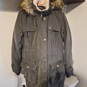 May include: A dark olive green parka jacket featuring a hood with a faux fur trim, a cream-coloured fleece lining, a front zip closure with a snap-button placket, two chest pockets, two large lower flap pockets, and ribbed knit sleeve cuffs.