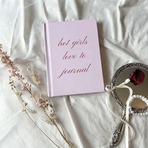 Może przedstawiać: Różowy dziennik z napisem "hot girls love to journal" w czerwonych literach. Dziennik leży na białym materiale z srebrnym lusterkiem ręcznym i suchym bukietem kwiatów.
