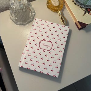 Może przedstawiać: Biały dziennik z wzorem czerwonych wiśni i napisem "Diary" w ozdobnej ramce. Dziennik leży na białej powierzchni, obok butelki perfum, lusterka i książki.