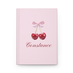 Può includere: Un diario rosa con un'illustrazione ad acquerello di due ciliegie e un fiocco rosa. La parola "Constance" è scritta in corsivo sotto le ciliegie. Il diario ha un design pulito e minimalista.