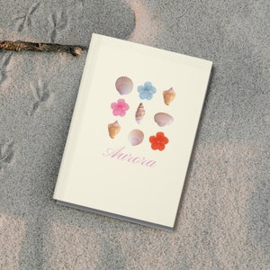 May include: A white notebook with a seashell and flower design. The notebook has the word "Aurora" written on the front in pink.