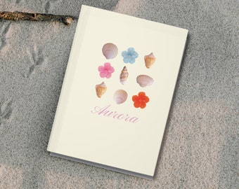 Può includere: Un quaderno bianco con un design a conchiglie e fiori. Il quaderno ha la parola "Aurora" scritta in rosa sulla copertina.