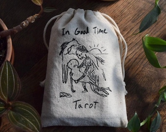 May include: A beige drawstring pouch made of canvas fabric featuring a black ink illustration of a skeleton embracing a figure, a horse, and a sun. The text "In Good Time" is printed above the illustration, and the word "Tarot" is printed below.