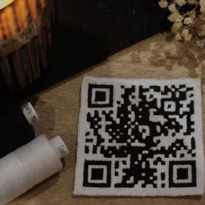 May include: A white square coaster with a black and white QR code printed on it. The coaster is lying on a wooden surface.