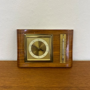 May include: A wooden framed barometer with a round analog dial and a thermometer on the right side. The dial has a black needle and white numbers from 70 to 80. The thermometer has a red needle and Celsius markings.