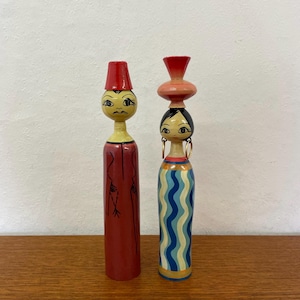 May include: Two vintage wooden dolls with painted faces and outfits. One doll wears a red fez and a red robe. The other doll has a black bob, gold earrings, and a blue and white wavy patterned dress. The dolls are decorative.