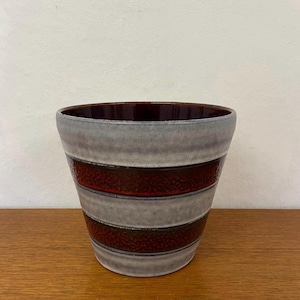 Pode incluir: Um vaso de cerâmica com um design listrado cinza e bordô. O vaso tem formato cilíndrico com interior marrom escuro. As listras são horizontais e uniformemente espaçadas ao redor do exterior. O vaso está sobre uma superfície de madeira.