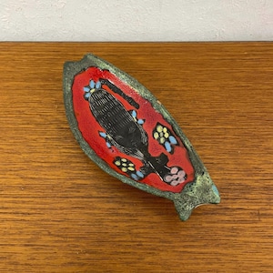 May include: A ceramic dish shaped like a fish with a red, green and black glaze. The dish is decorated with a stylised figure and colourful flowers.