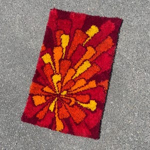Op de afbeelding: Een rechthoekig vloerkleed met een retro bloemenpatroon. De dieprode achtergrond contrasteert met een uitbarsting van oranje en gele kleuren die uit het midden komen. Het vloerkleed heeft een zachte, pluizige textuur.