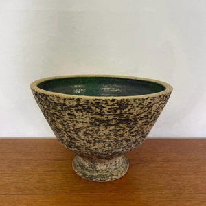 May include: A speckled ceramic bowl with a dark green interior. The bowl has a wide, flared shape and sits on a short, pedestal base. The exterior has a textured, speckled pattern in shades of brown and black.