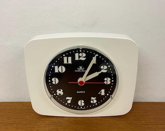 May include: A white, square-shaped quartz alarm clock with a black face. The clock face features white numerals, white hands, and the text "MEISTER ANKER QUARTZ". The clock has a red second hand.