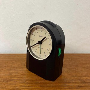 Puede incluir: Un despertador Garant negro con radio control, con esfera color crema y manecillas negras. El reloj tiene un botón verde en el lateral y muestra la hora con números negros. El reloj está sobre una superficie de madera.