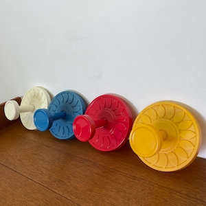 Puede incluir: Cuatro perchas de pared coloridas en fila. Los ganchos son blancos, azules, rojos y amarillos. Cada gancho tiene una base circular con un patrón decorativo y un pomo cilíndrico. Los ganchos están montados en una pared blanca y un suelo de madera.